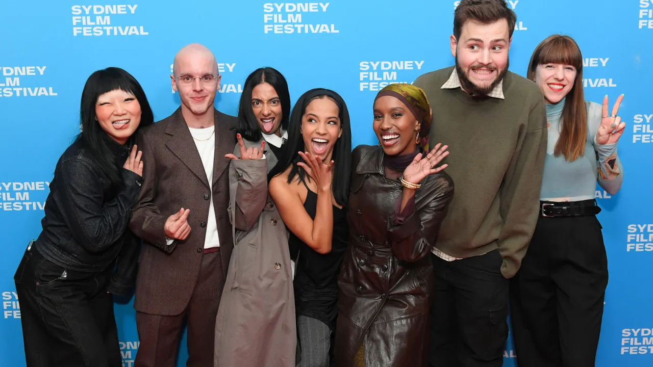 A group of seven people pose playfully on the red carpet at the Sydney Film Festival. They stand in front of a bright blue backdrop with the festival’s logo repeated across it. Each person expresses joy and excitement, smiling and making fun gestures—such as peace signs, hand horns, and exaggerated faces.