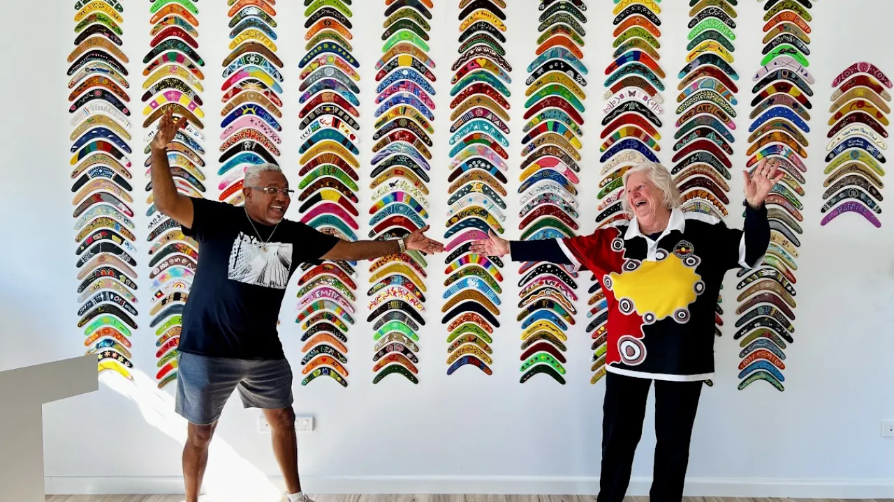 A man and a lady dance in front of 378 painted boomerangs displayed on a gallery wall at the Opening of the WWCACC Community Boomerang Wall Project.