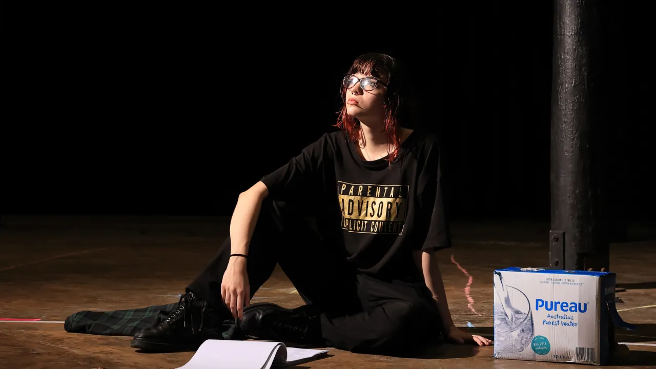 A performer from Rebus Theatre’s Tent City sits on the floor of a darkened performance space, illuminated by a warm spotlight. Surrounded by minimal props, the image captures a quiet, reflective moment from the production’s creative development.A performer wearing black sits on the floor of a dark theatre under a spotlight, with papers and a cardboard water box beside them.