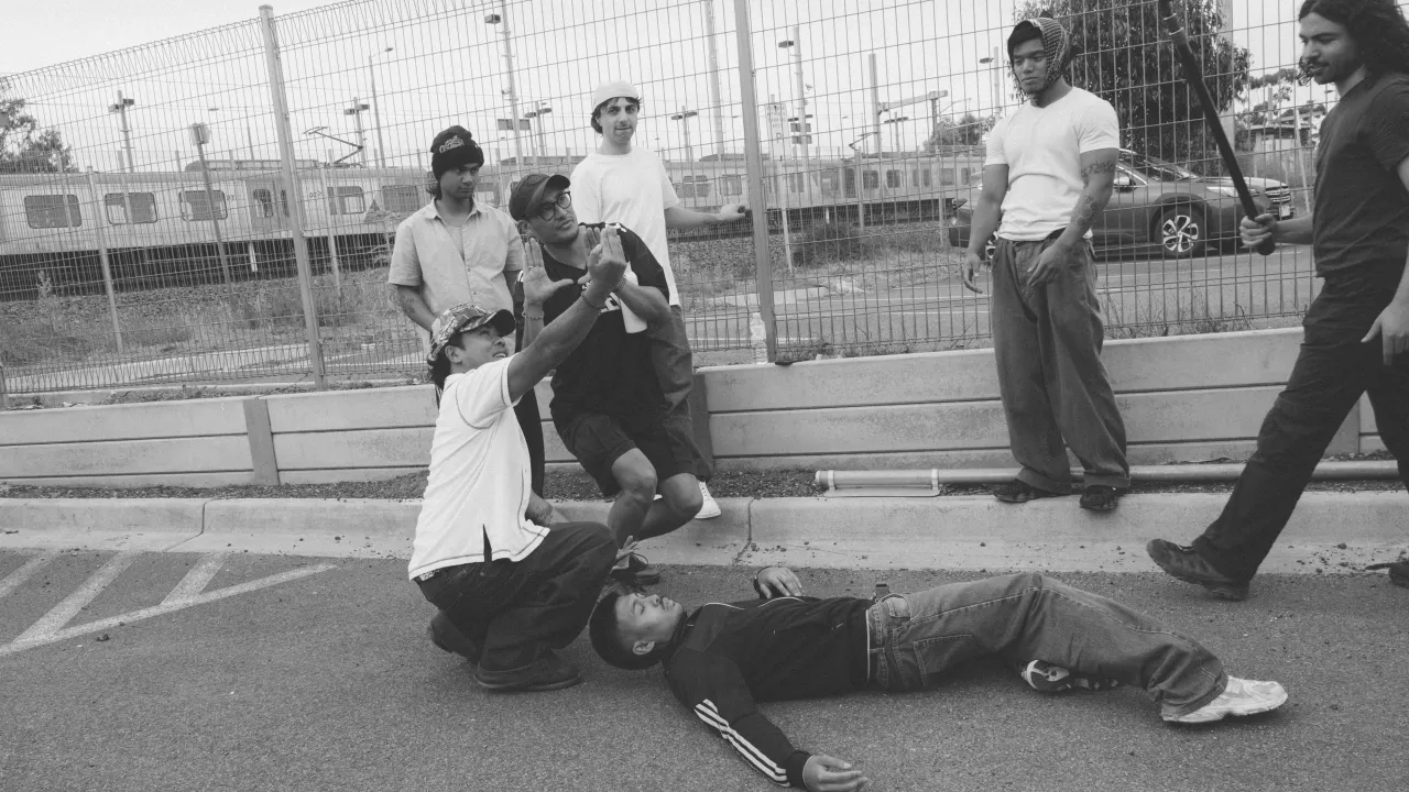 A director and cinematographer stand among a group of actors preparing to shoot a scene for a film. One actor lies on the ground as the director points into the distance, his gaze follows. The cinematographer crouches behind raising his hands to make a frame - through which he assesses the scene.