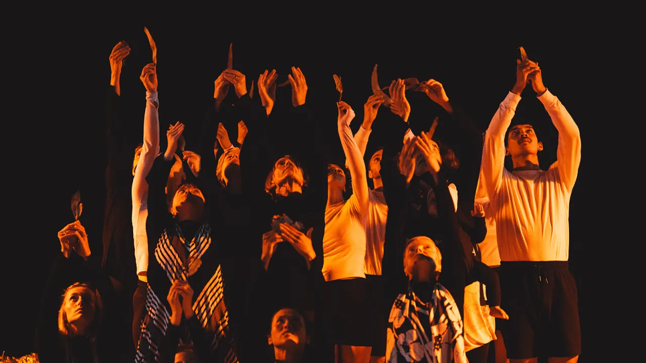 A group of dancers are pictured in yellow/orange lighting with their arms held above their heads