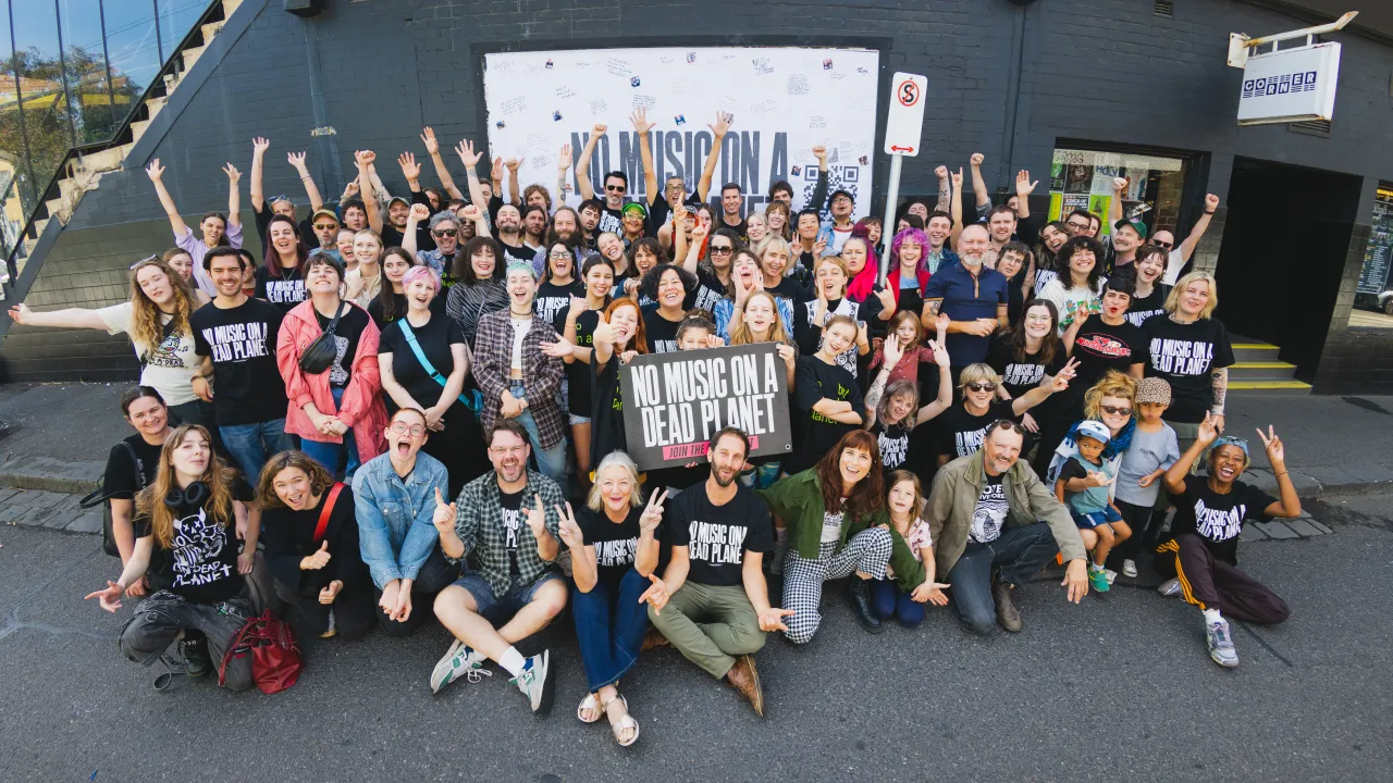 A large group of people pose outside the Corner Hotel, many raising their hands and smiling. They hold a large sign reading 'NO MUSIC ON A DEAD PLANET' and several people wear matching T-shirts with the same message.