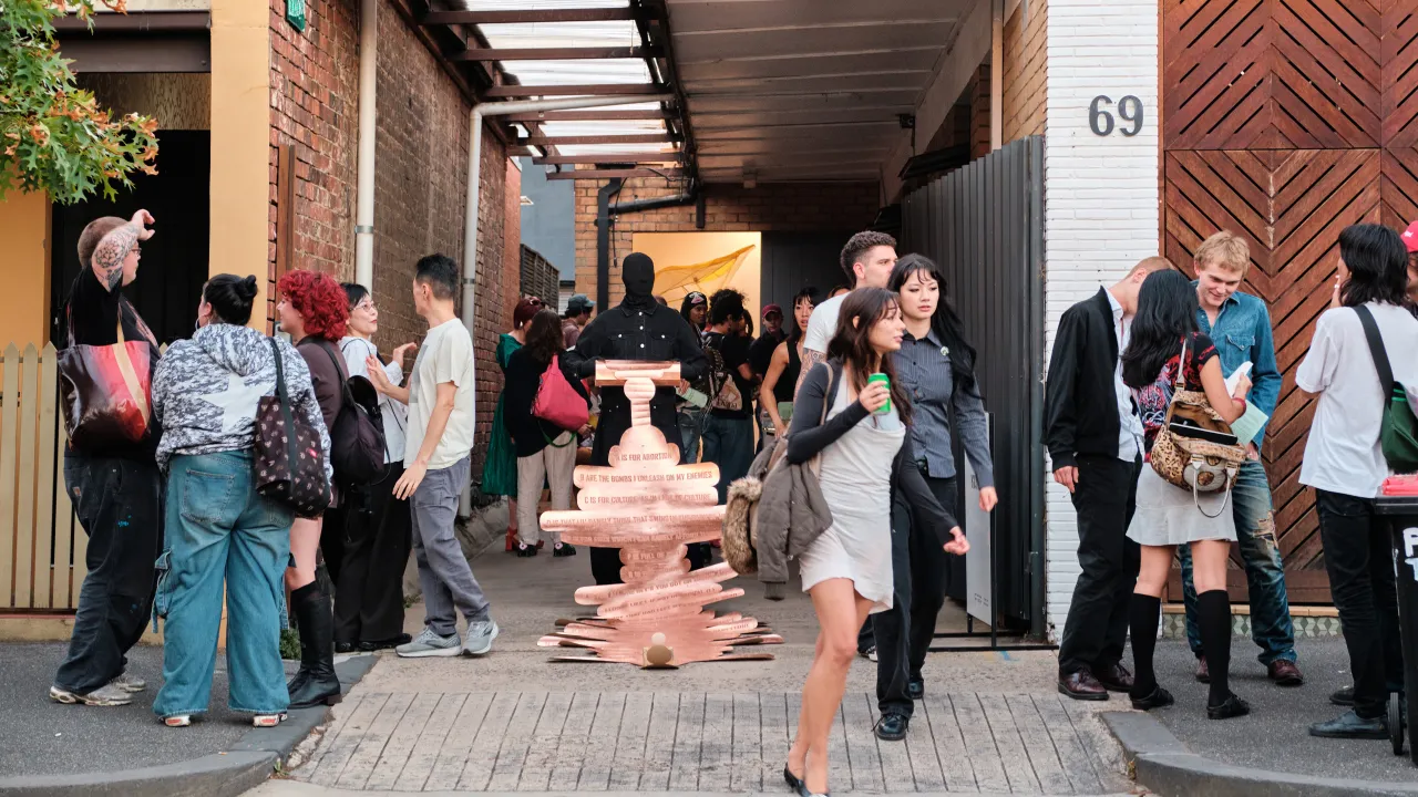 A photograph of Thang Do’s performance, outside KINGS Artist-Run. Do is dressed in all black, with a mask of stretch fabric obscuring his face. He holds a long unfurling roll cut from card, with rounded bubble-like rows divided by horizontal creases. The surface is painted with bronze metallic paint, and each row has hand-written text in sans-serif all-caps. Do stands still, surrounded by an energetic crowd conversing and walking. In the far background, a sliver of the interior gallery is visible.