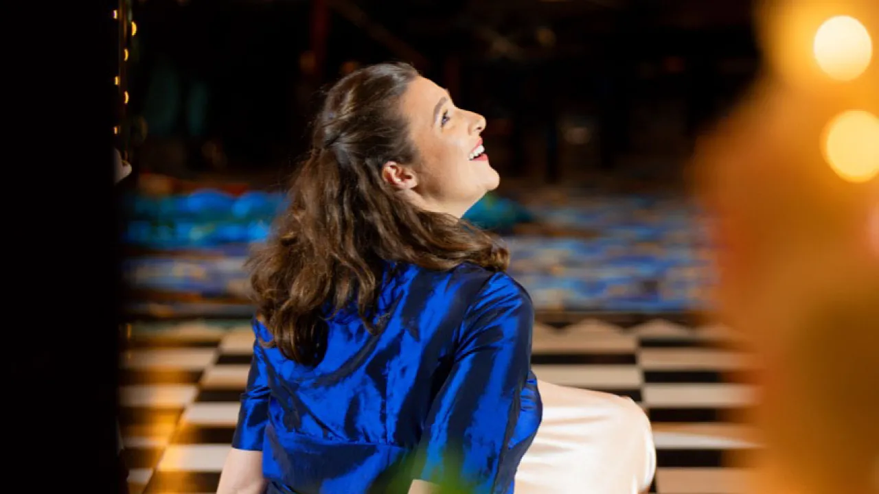 Performer seated on a black-and-white checkered floor during a staged performance, wearing a vivid blue satin top and cream skirt. Warm lanterns and string lights frame the scene, while musicians appear softly out of focus in the background, creating an intimate theatrical atmosphere.