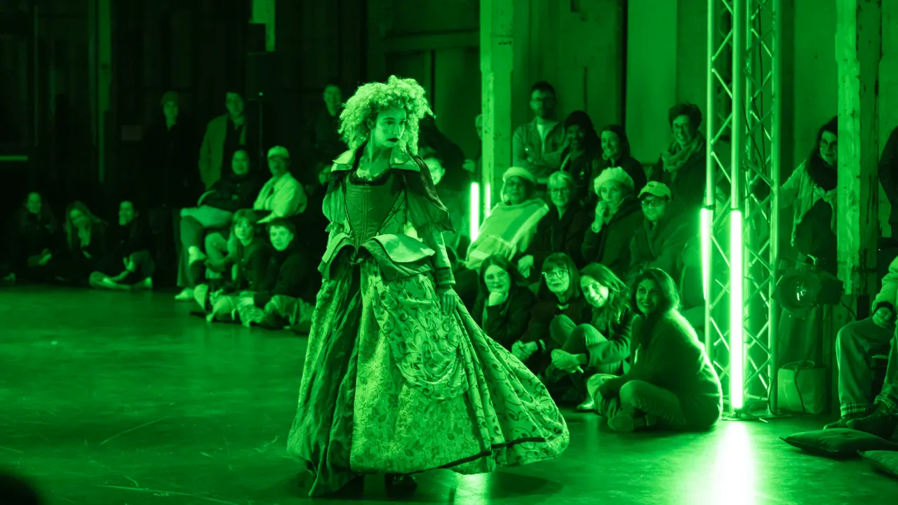 Bathed in bright green light, a dancer in a corseted period-style gown, curly wig, and clown-like makeup performs in front of a small seated audience