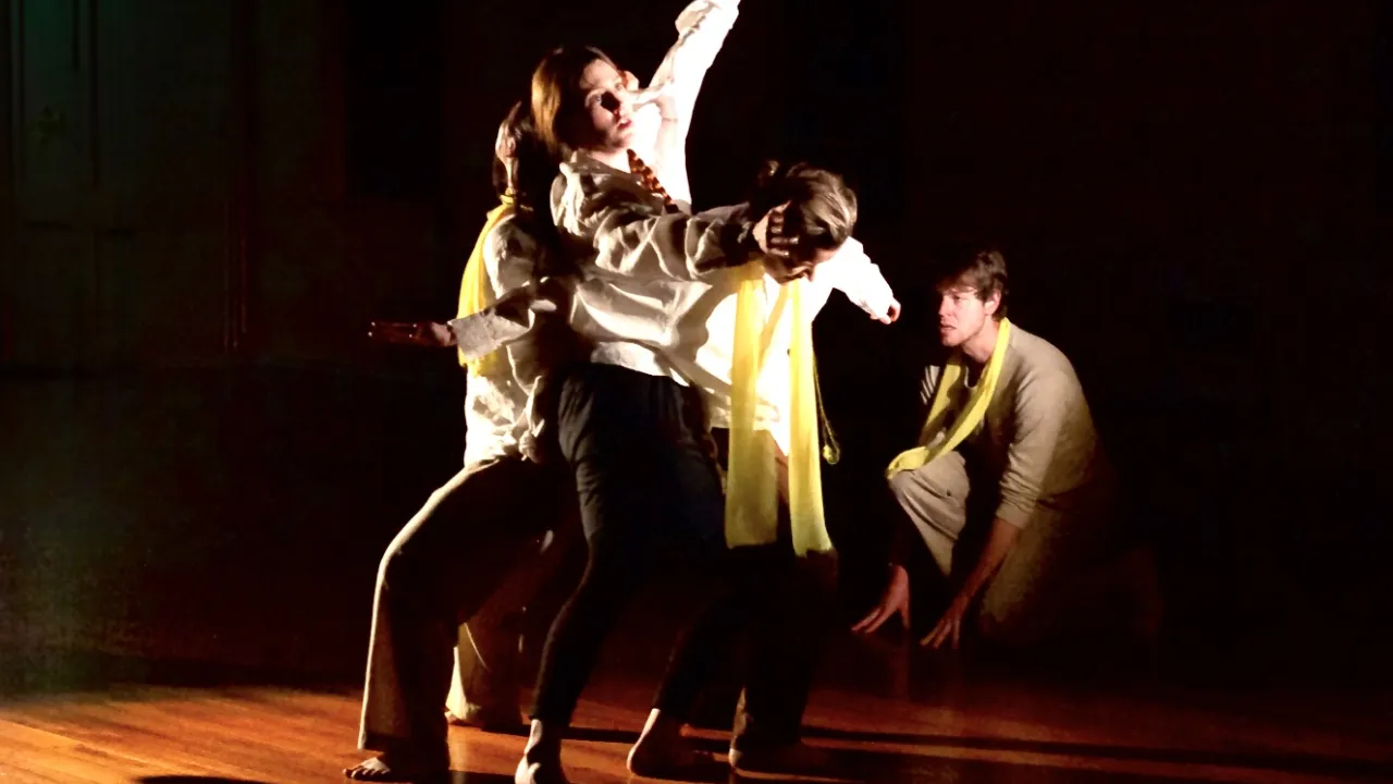 A group of four performers engages in a dramatic, physical theatre performance on a wooden stage bathed in warm, angular stage lighting against a dark background.