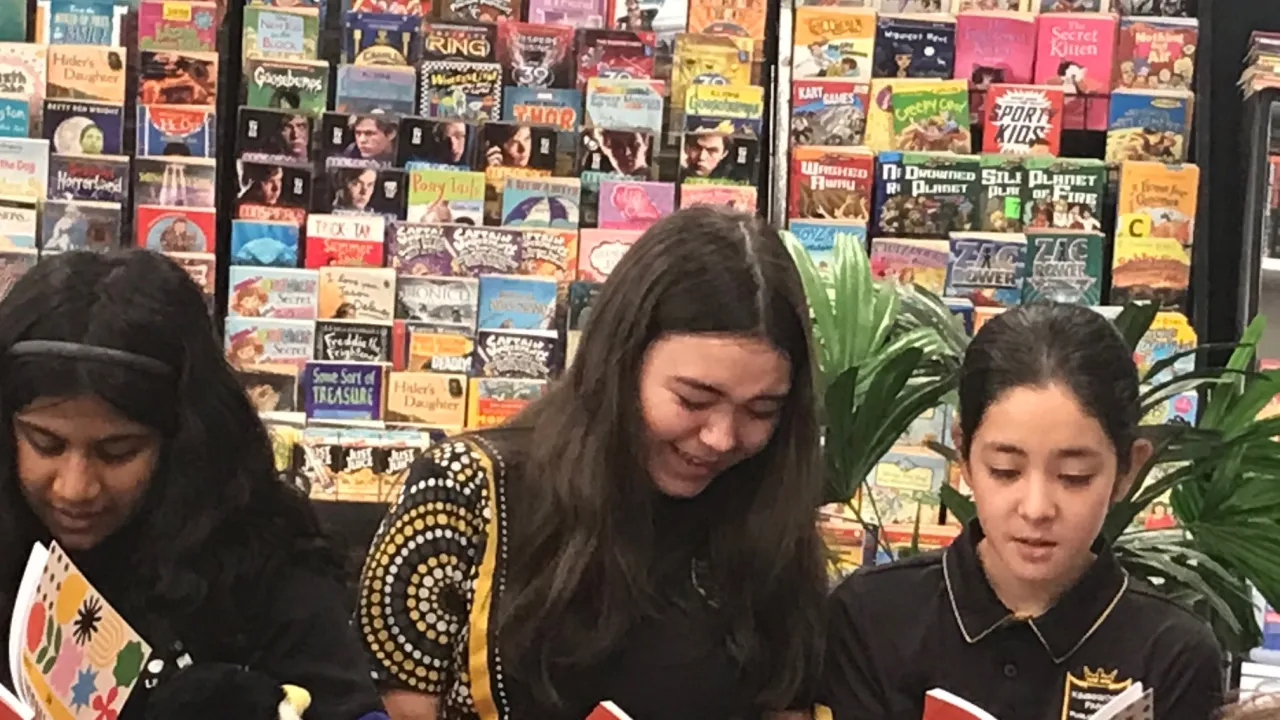Young students read their own published work in front of library shelves full of books