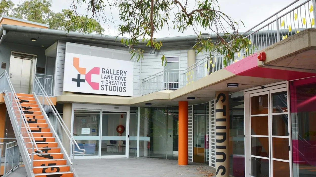The entrance to Gallery Lane Cove + Creative Studios in Lane Cove, Sydney. Bright orange stairs painted with the word "Gallery" lead to the upper level, while a column marked "Studios" stands beside glass doors to the ground-floor studios. The centre's orange, pink and black logo sits above the entry, framed by overhanging gum tree branches. 