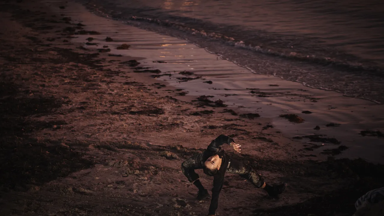 A dancer dressed in dark pants, top and shoes arches backward on a muddy, seaweed-strewn shoreline at dusk, one arm reaching up with fingers splayed, the other braced against the ground, lit warmly against dark rippling water. Their upside-down face looks directly at the camera.