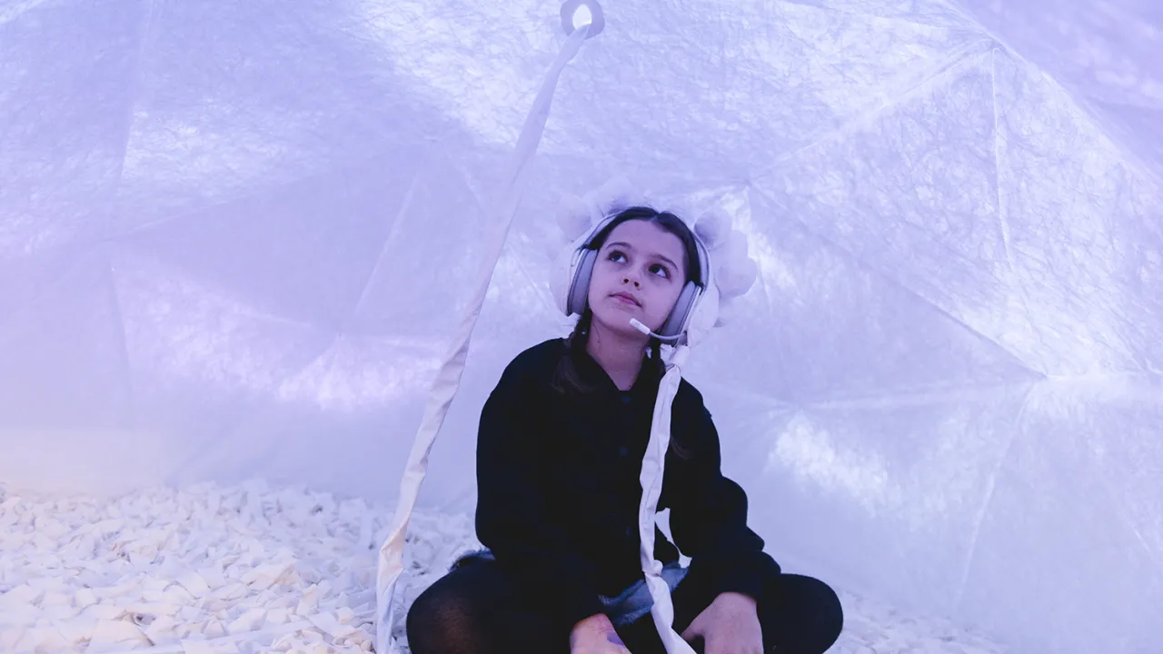 A child sits cross legged in a soft, white enclosed space, illuminated with lilac light. They wear embellished white headphones connected to the roof of the dome via a white cord.