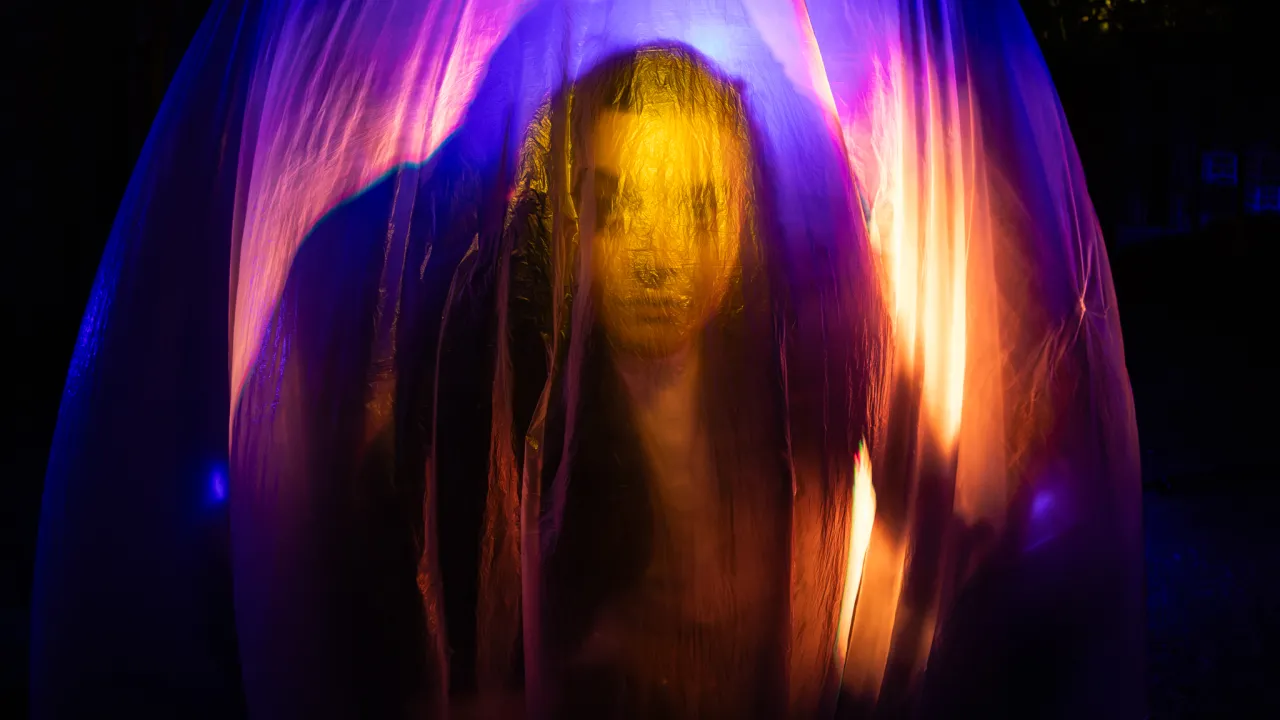 The silhouette and facial features of Cheryn Frost are just visible through a sheet of material. Cheryn is lit from behind with purple and orange light, casting an ethereal shadow that frames Cheryn’s torso. 