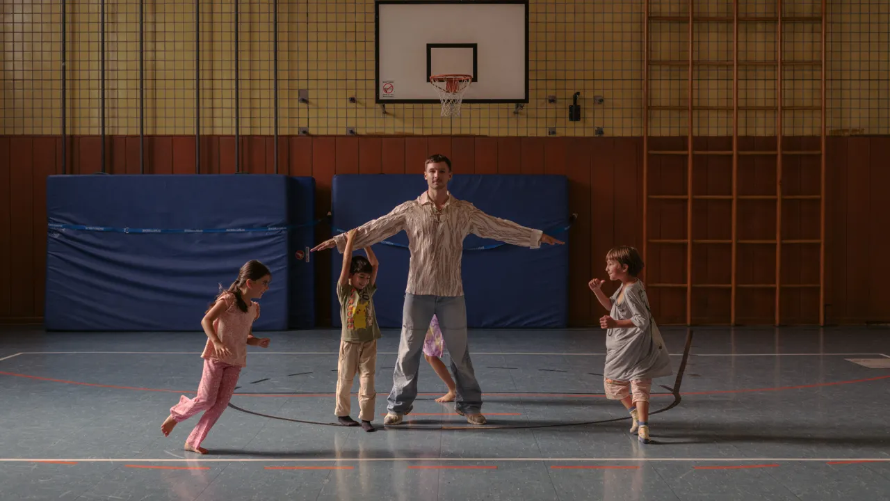 A dancer stands with their arms outstretched in the centre of a school gymnasium while four young children move playfully around them. Blue gym mats, a basketball hoop, and wooden climbing frames frame the scene, contrasting the dancer's stillness with the children's lively energy.