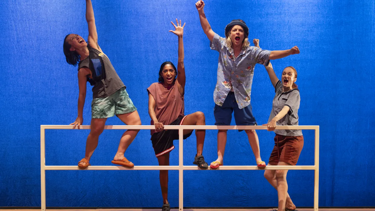 Performers stand together on a dramatically lit stage, arms raised and holding red poles, capturing the energy, boldness and collective spirit of live theatre.