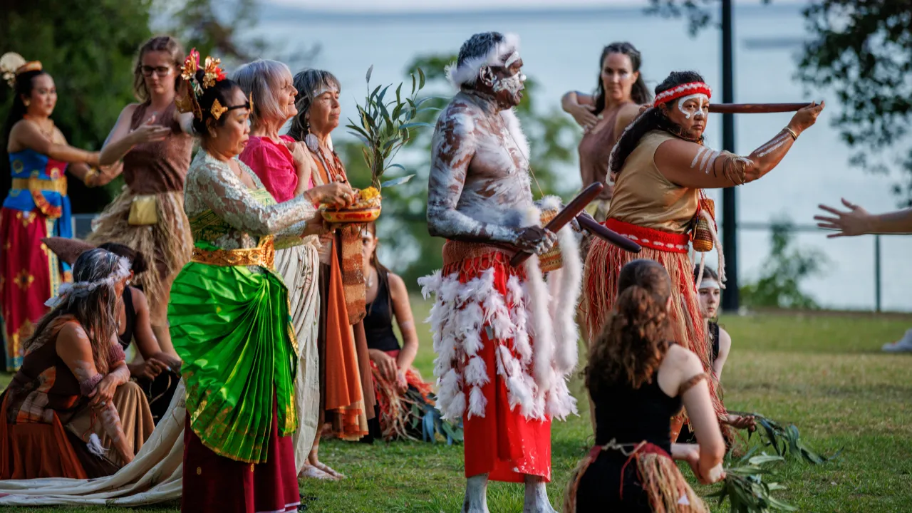 Antidote 2025. Photo of the performance featuring Yolŋu, Larrakia, and Balinese performers. The perfomers are on grass, surrounded by trees. You can see the ocean in the background.