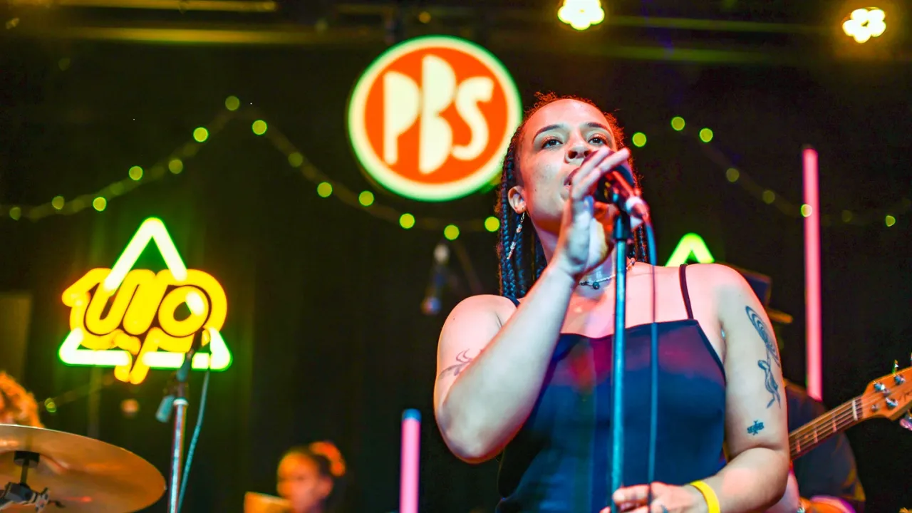 Hero image description: Owelu Dreamhouse performing live at CY Venue as part of PBS Drive Live 2026. The image features the singer, Nkechi, in focus singing into a microphone, with blurred band members in the background. There is a light of the PBS logo to the left of Nkechi's head, and a neon Up Bank logo further to the left of screen.  Alt image text: A woman on stage singing into a microphone