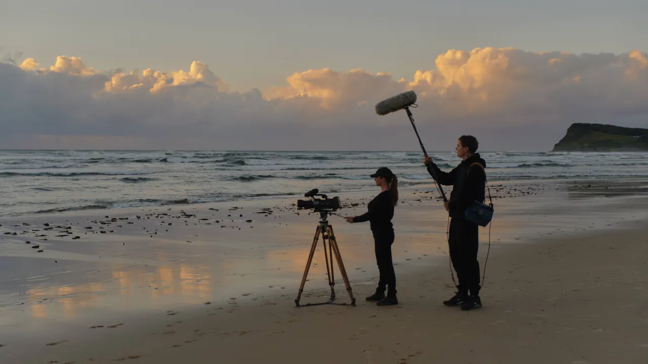 Two people film with a camera and boom microphone on a sandy beach at sunrise, facing the ocean with clouds in the sky.