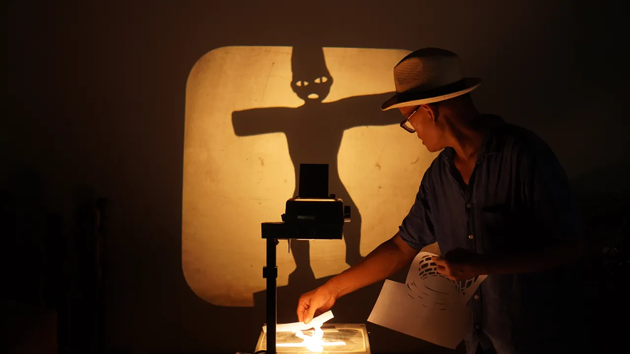 A man wearing a hat leans over an overhead projector holding a wayang (puppet) in the shape of a person with cutouts for the eyes. The overhead projection makes the puppet larger, taking up the centre third of the projected wall. 