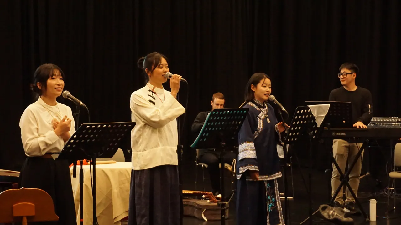 Three performers sing into microphones during a Gold Mountain Wives development showing, accompanied by musicians on keyboard and other instruments.