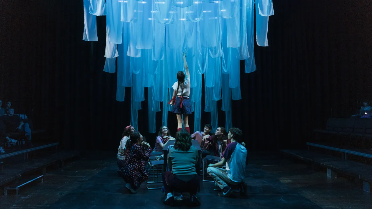 A young girl is standing on a table with her aim reaching up into a waved curtain of fabric that is coloured blue. A group of young people crouch on the ground looking up at her. 