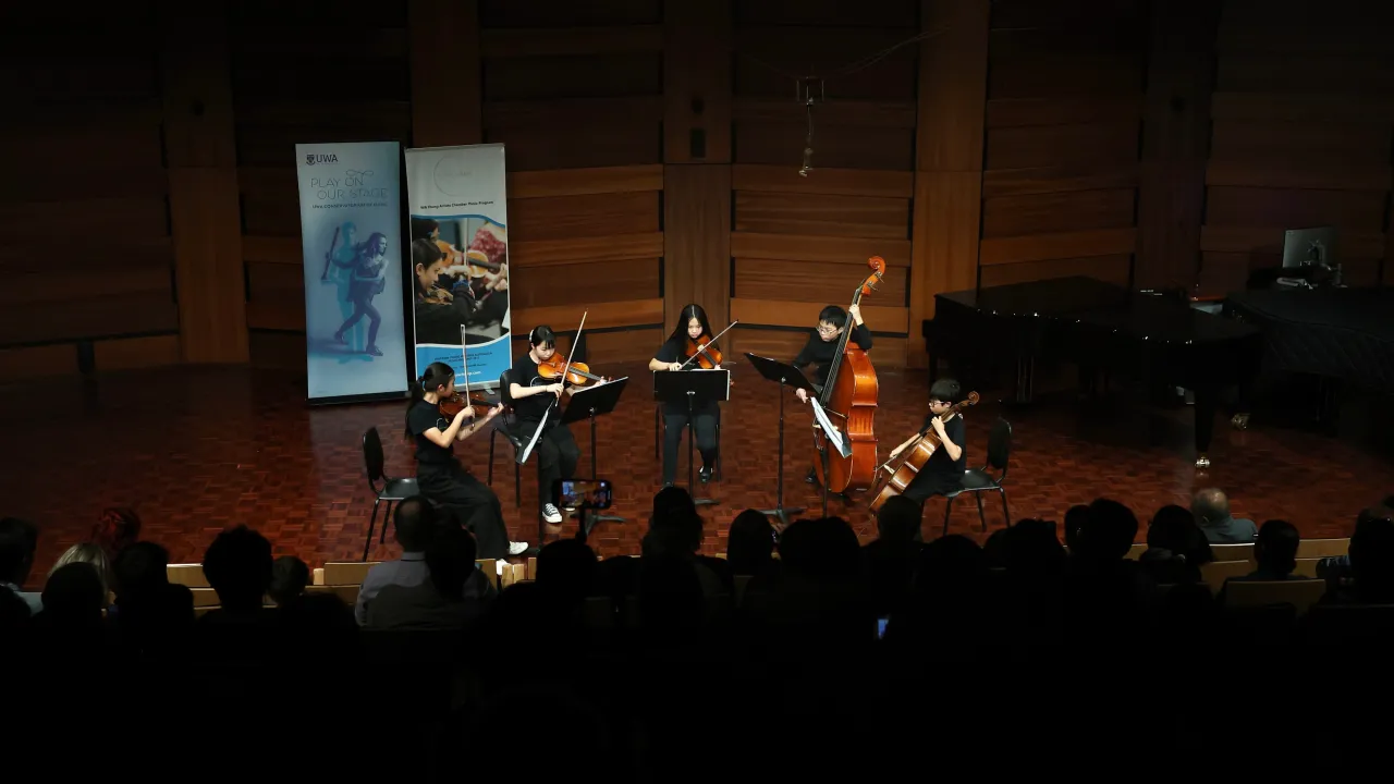 Image depicts a group of five young people playing string instruments in a formal concert setting in front of an audience. In the background are a grand piano and banners.