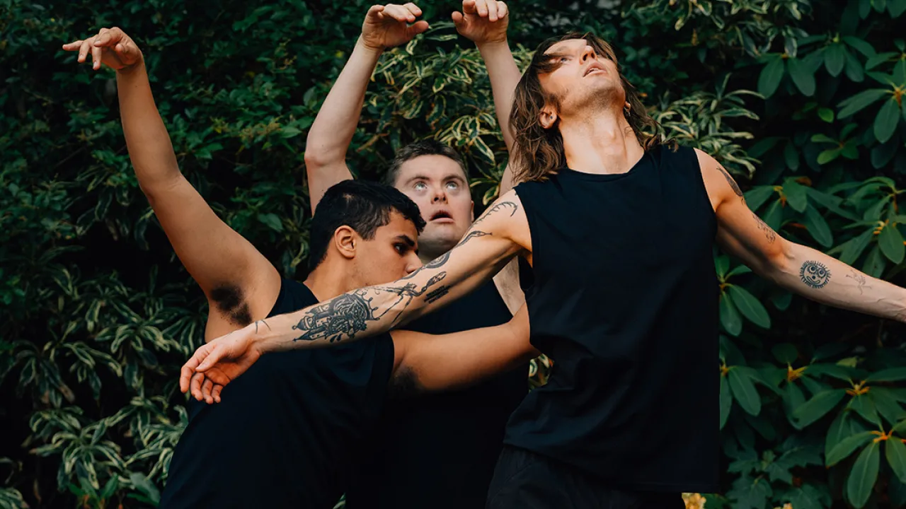 Three dancers, wearing black costumes, in front of a lush green backdrop