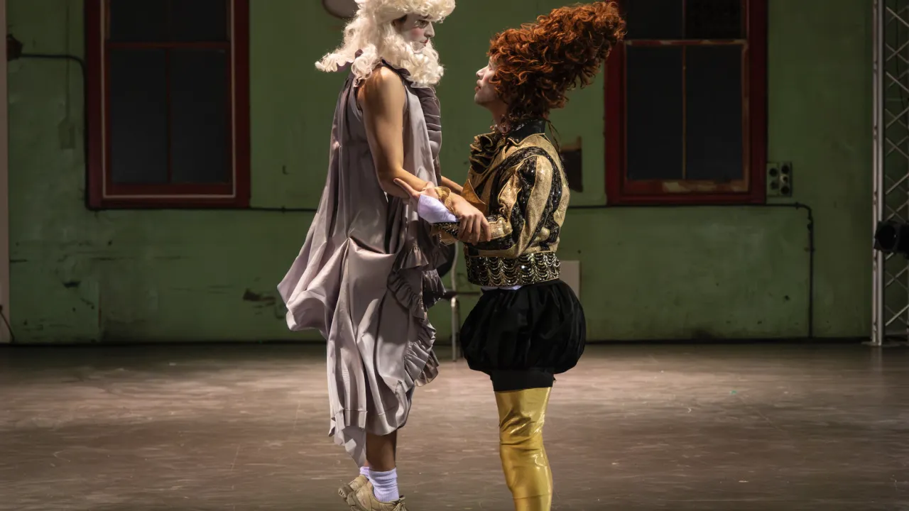 One dancer hovers vertically above the ground, held by another dancer. Both are dressed in colourful medival costumes, with large wigs and clown-like make-up.