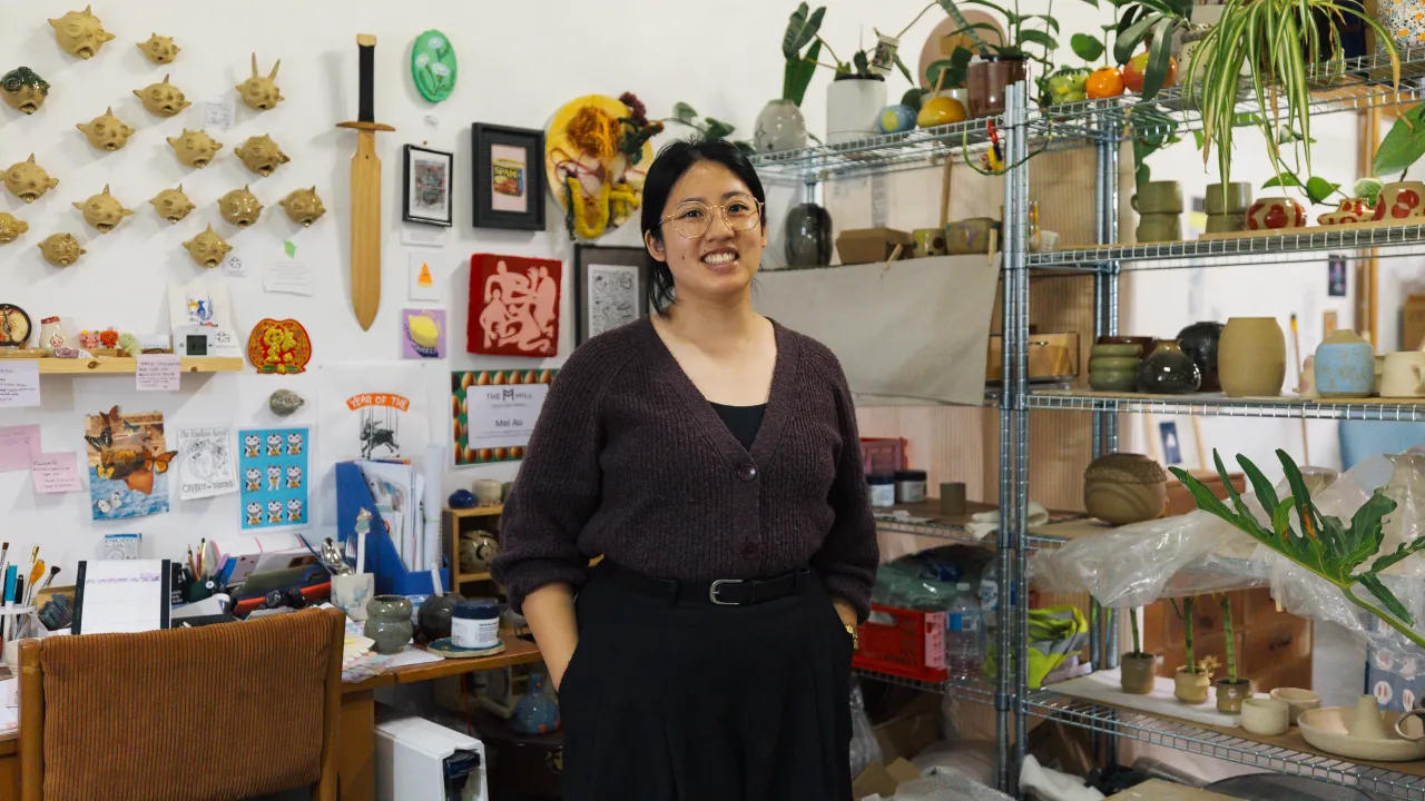 Ceramicist Mel stands in their studio, smiling broadly, behind them are shelves filled with ceramics and a wall filled with ceramic objects.