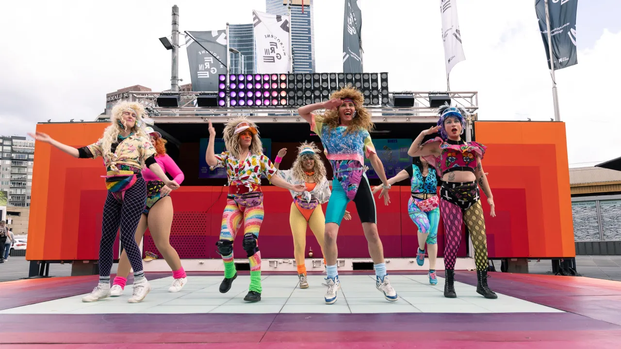 Performers in colourful activewear dance on an outdoor stage, with Melbourne Fringe flags and city buildings behind them.