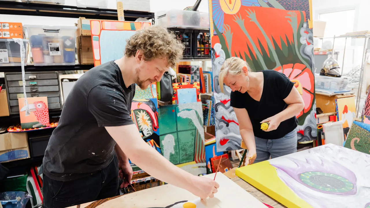 A photograph of two artists in a messy, brightly coloured artist studio. The artists are both bent over a table, painting on separate canvasses.