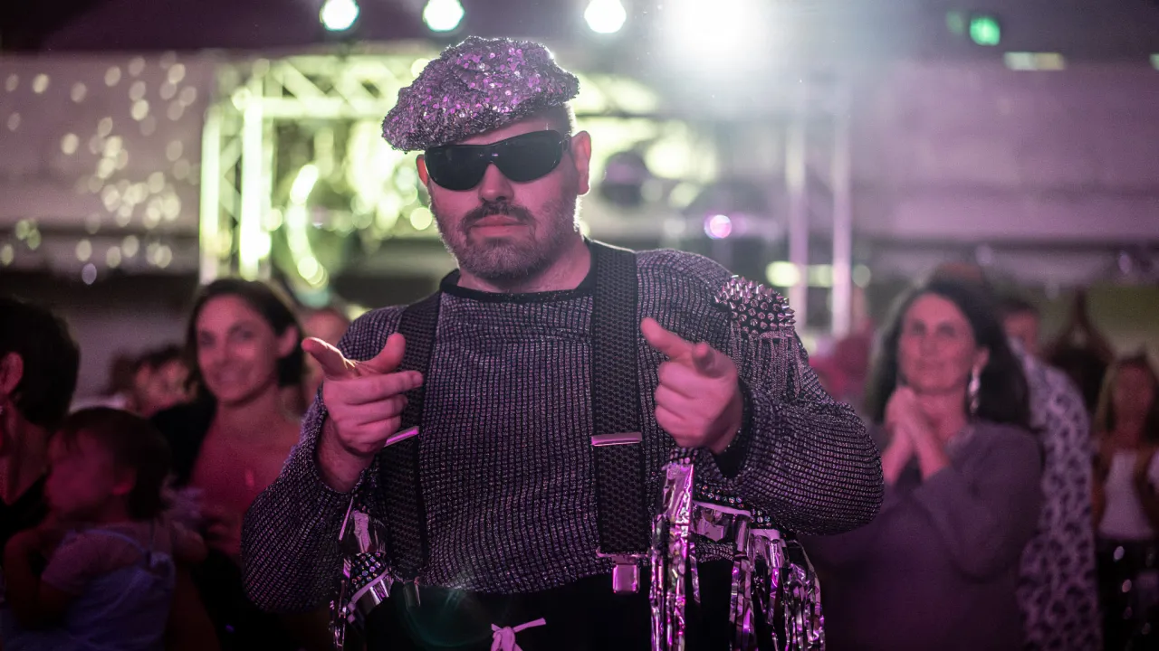 A person wearing a silver sequined cap, dark sunglasses and a shimmering silver outfit points playfully toward the camera while standing in front of a lively crowd at a disco event. Bright stage lights and a blurred audience create an energetic atmosphere, with metallic costume details reflecting the colourful lighting.