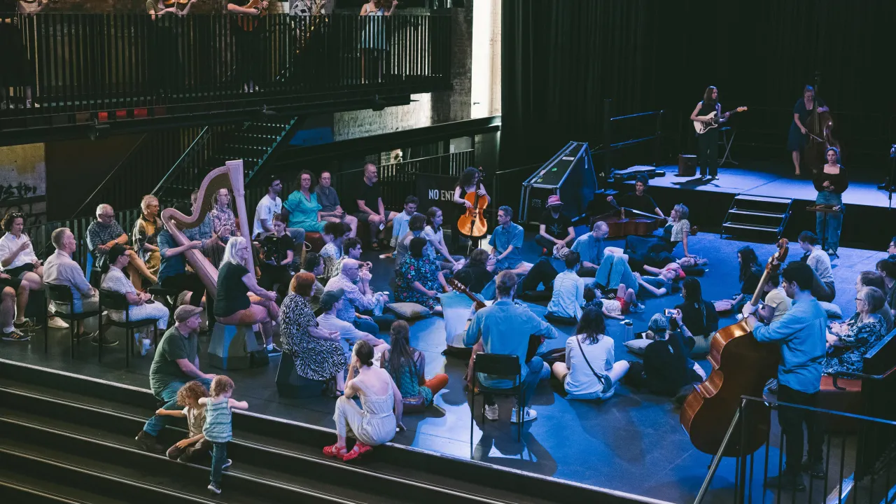 A photograph of "Dots+Loops MEDITATIONS", Sunday 1 December 2024 at Brisbane Powerhouse. A large ensemble of musicians are spread throughout the Turbine Hall of the Powerhouse, some in amongst the audience seated on cushions in the centre of the room, some musicians on stage, and some on the balconies surrounding the room.