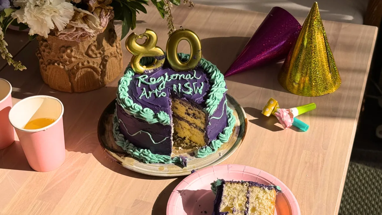 Image description: A dark purple and eucalyptus green birthday cake sits in the centre of a wooden table in bright natural light. The cake is cut with one slice taken out, and is decorated with the words, ‘Regional Arts NSW’ written in icing and burnt gold candles saying the number 80. Surrounding the cake is table set for a birthday party with a vase of brightly coloured flowers, pink paper cups, two pointy party hats and a paper plate with a slice of the cake on it.  Alt image text: Birthday cake with t