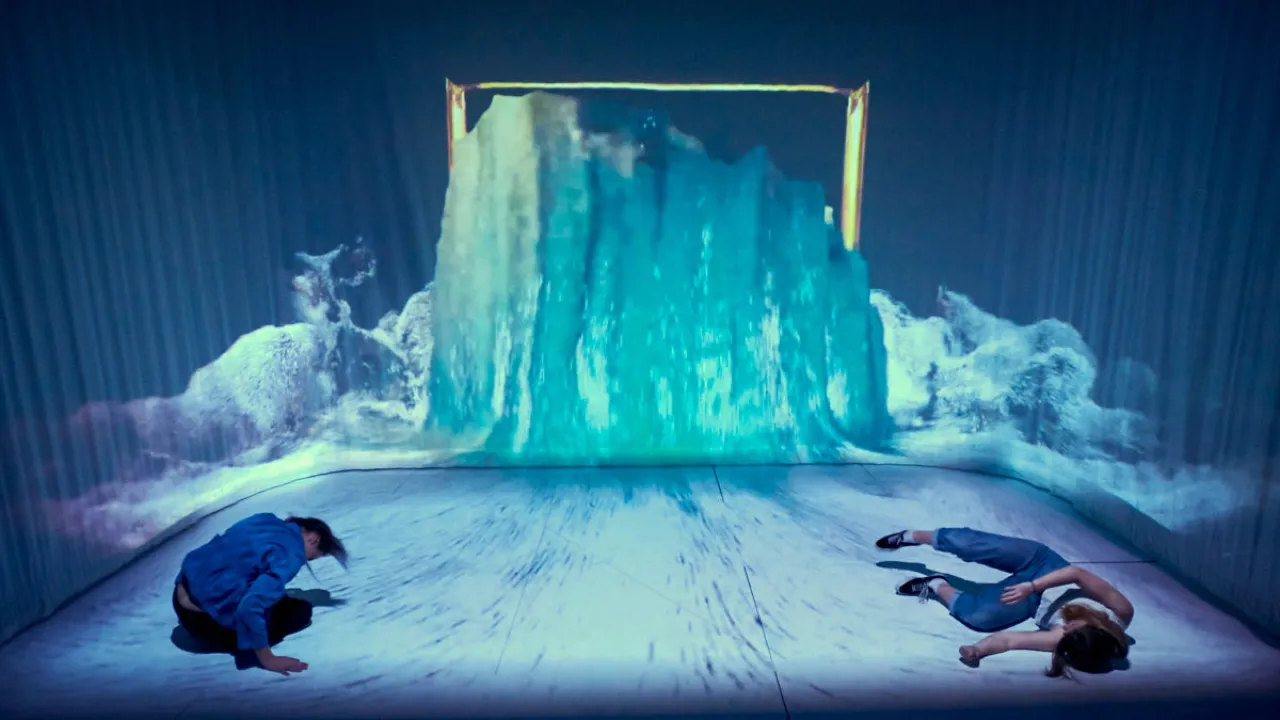 The image captures a striking moment from 'Secret of the Raft', where water appears to burst through the frame of a painting, as though the image has broken free into the physical world. Two performers lie immersed in the cascading digital landscape, blurring the boundary between the painting, the stage, and reality. The scene evokes a world shaped by memory, danger, and survival.