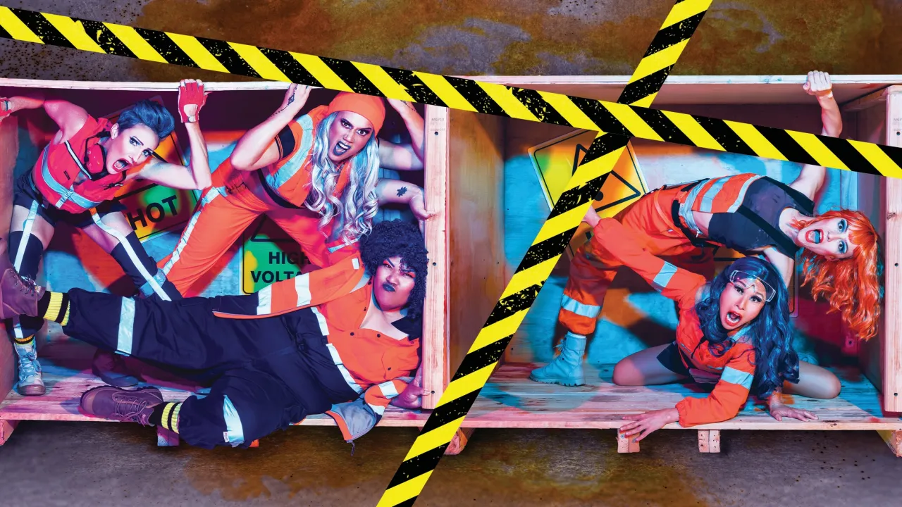 Promotional photo for Polytoxic's 'Dangerous Goods': five performers in orange and navy hi-vis safety wear, gloves, and colorful wigs pose dramatically inside a wooden shipping crate, mouths open in exaggerated expressions. The crate's interior is painted with hazard signage reading 'HOT' and 'HIGH VOLTAGE.' Black-and-yellow diagonal hazard tape crosses the image, set against a rusted, weathered backdrop.