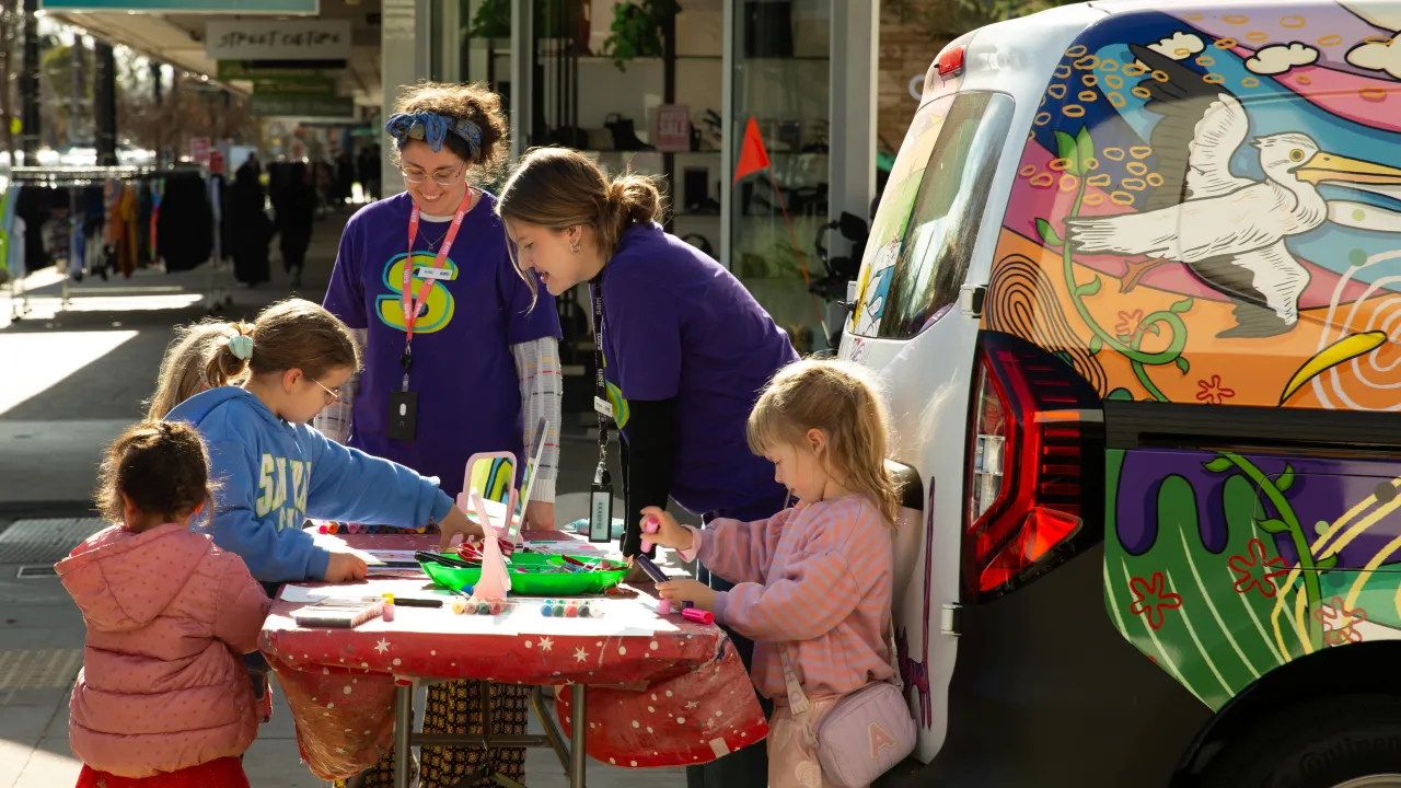 Two women help three young children with art activities beside a colourfully-decorated van in a public square.