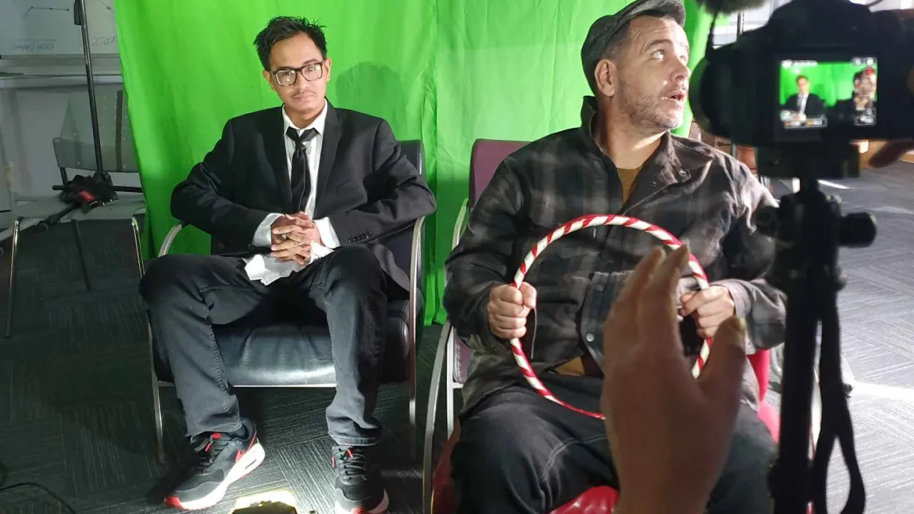 Behind-the-scenes StoryLab in action: two men sit before a green screen, one in a suit and one holding a steering wheel, filmed by camera crew.