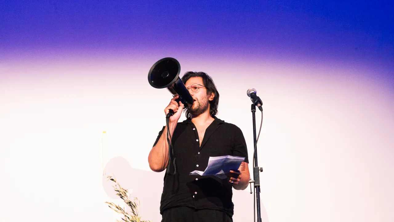 Diego Ramirez an artist with short dark brown hair and glasses and a black button-up short sleeve shirt, holding in his righthand a black megaphone to his mouth and speaking. In his left hand is printed paper. A microphone on a stand is to his left, and a branch of Olive leaves in front of him. 