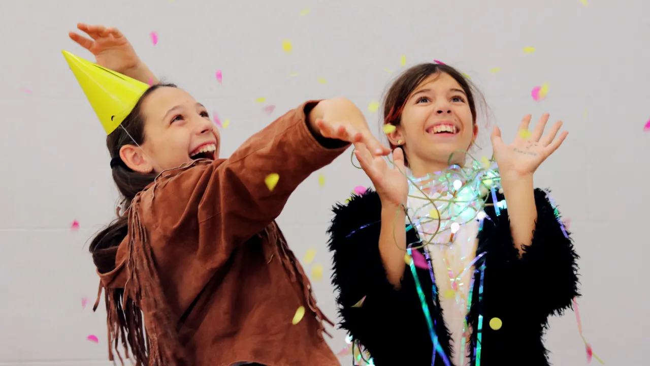 A photo of two young people smiling and looking up with arms out to catch the pink and yellow confetti is falling down around them. One is wearing a yellow party hat.