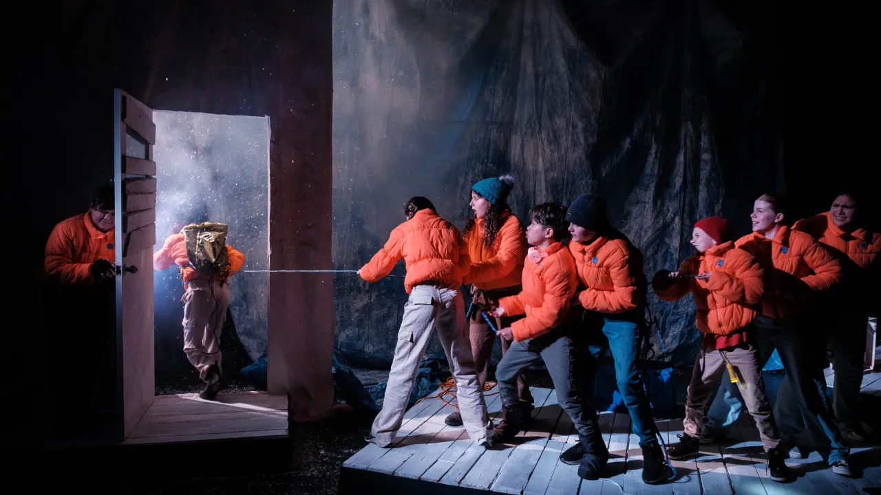 9 young performers wearing orange puffer jackets on a theatre set pulling a role through a doorway