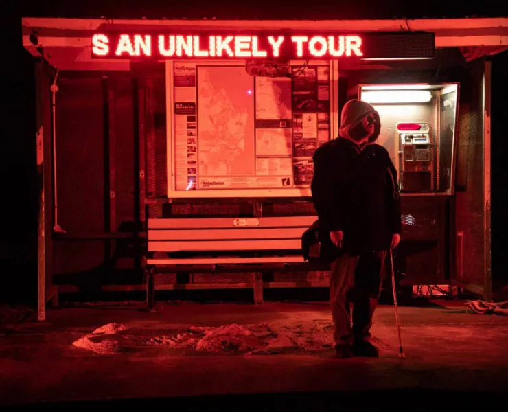 Man in hoodie with white cane in front of phone booth with neon sign and red colour wash