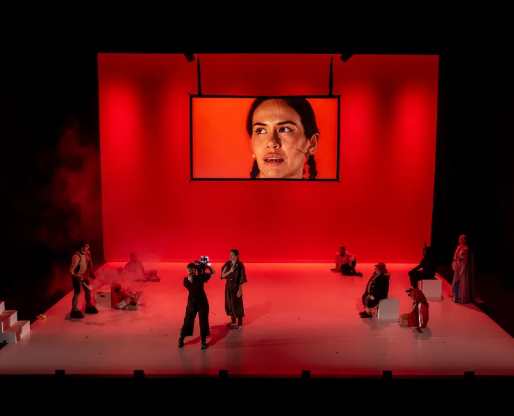 A photo of a performance on a large stage. The stage has a group of performers all dressed in black, spread across the stage. On the back of the stage is a screen that has a close up of a performers face. The stage is glowing red.