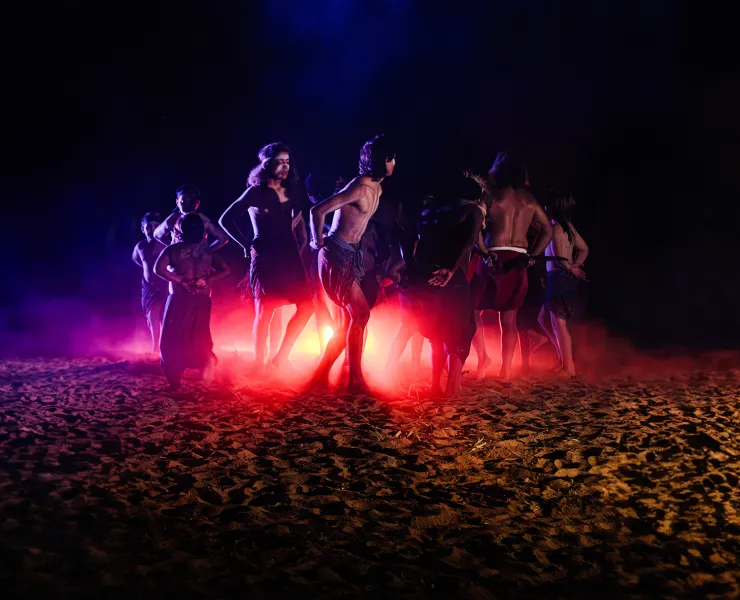 A photo of Indigenous dancers moving in a circle around a central red light. They are outdoors on sand under a dark sky.