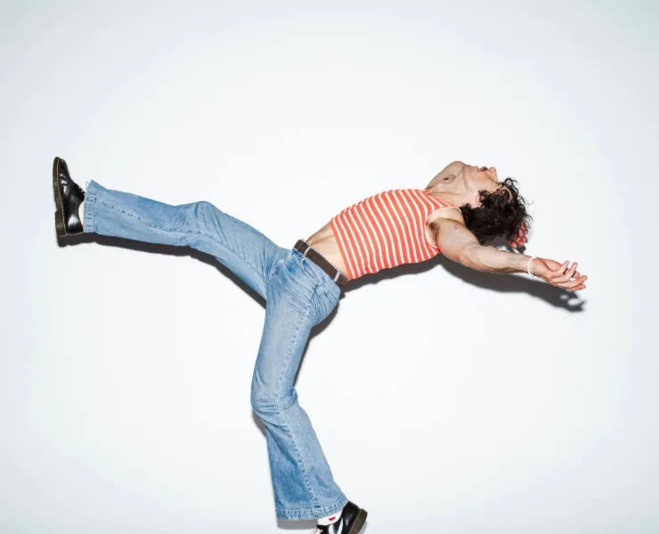 Go-Jo is wearing blue jeans and an orange and white striped tank top. He is kicking one leg up and opening up is arms.