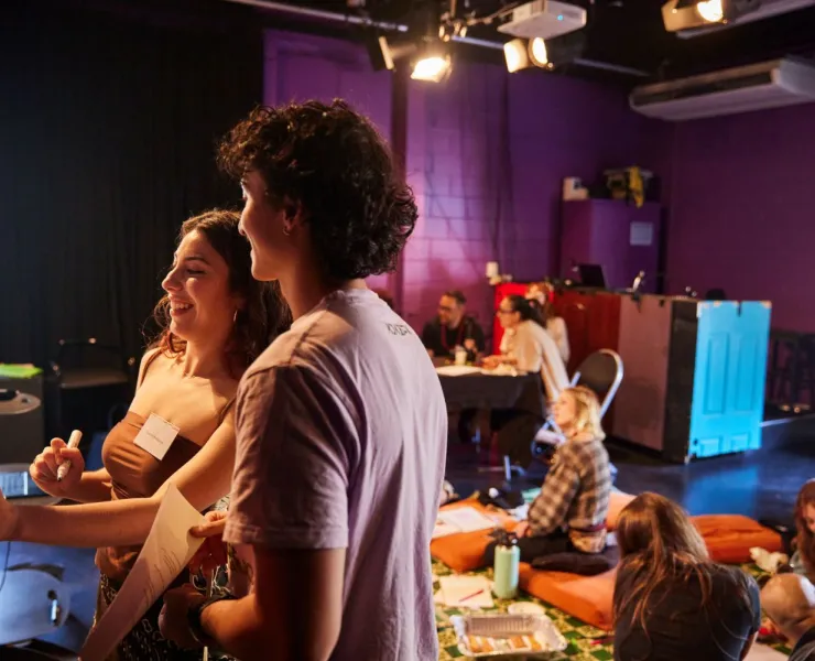A photo. Two people are working together at a white board, while there is a group working together, sitting down in a circle behind them.