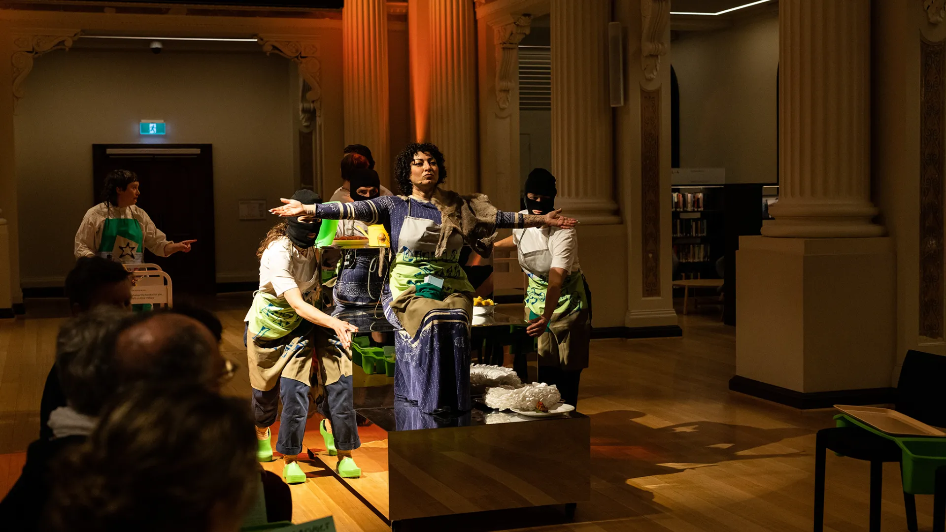 A performance with an audience watching. A central performer sits on a long podium with a number of other performers wearing balaclavas, pushing them forward.