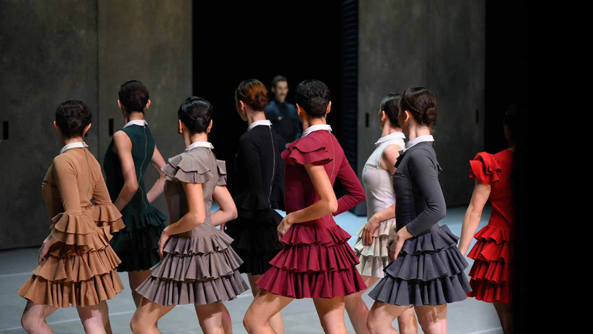 A photo of a group of dancers. They are all wearing matching dresses in different colours, deep red, greys, deep greens and light brown. They are all posing with their backs to the camera, and with one leg bent, hands on hips and heads all facing the same direction.