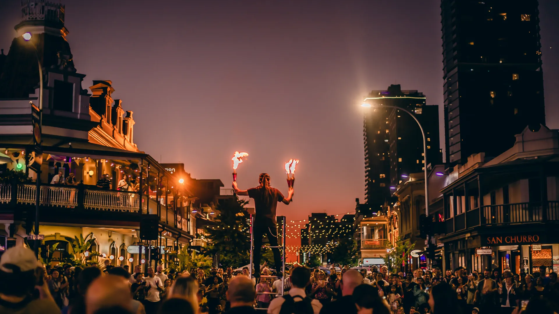 A photo of a large crowd in the middle of a city intersection. The large crowd watches as a street performer stands on the top of a lander, with multiple battons that are on fire in heir hands. The sky is as dusk, in a purple and orange ombre.