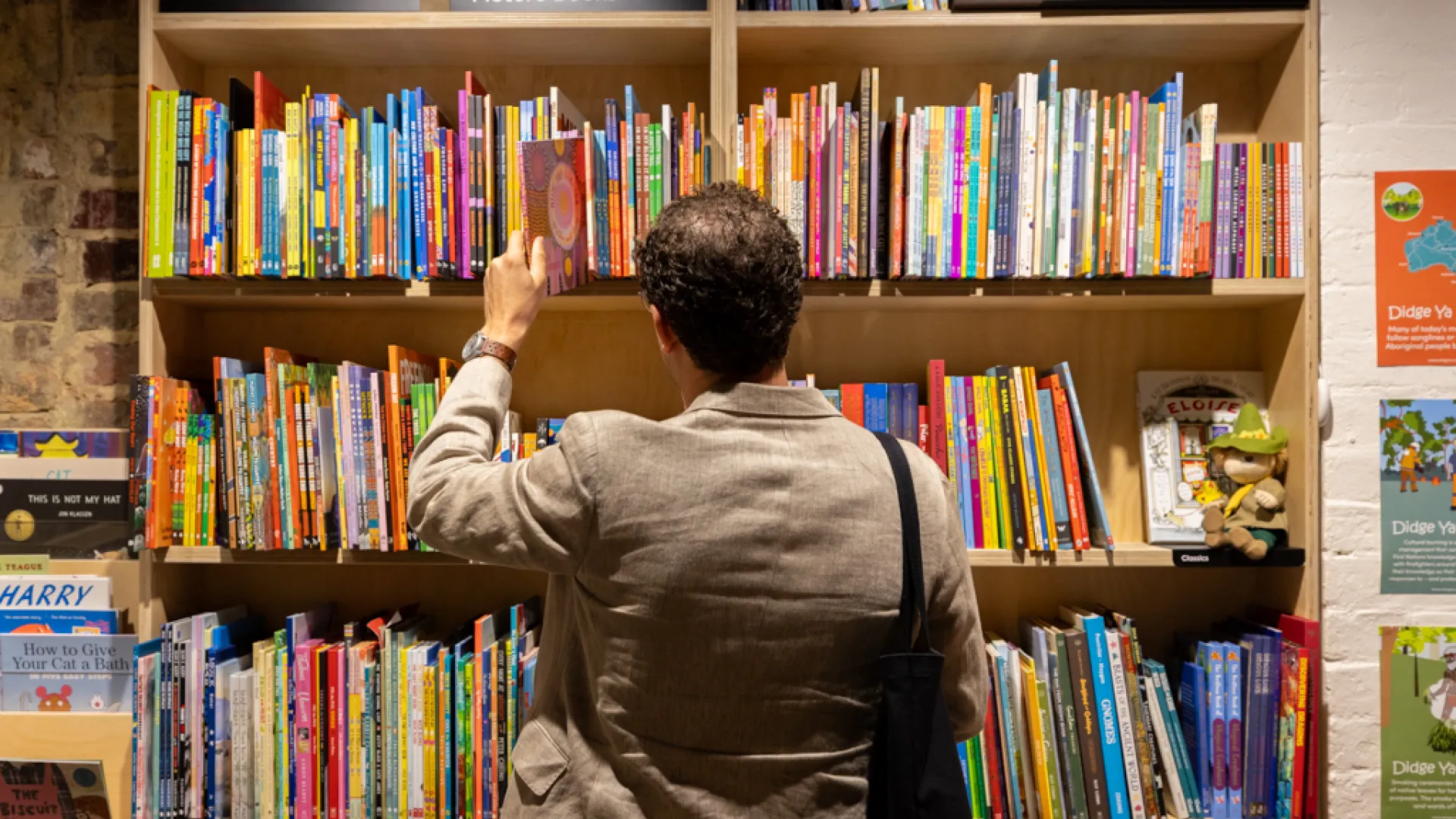 A man stands in front of a bookshelf full of childrens books, with his back to the camera. He is taking a book out from the top shelf. There are three sings that say 'First Nations', 'Australian Picture Books' and 'Children's classics'. 