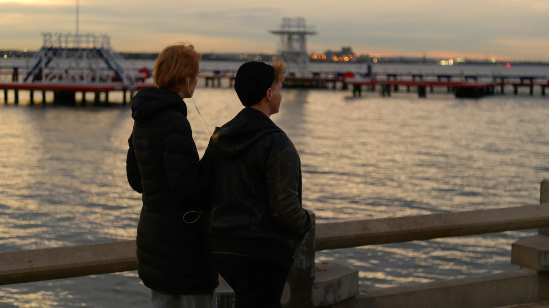 Two people standing at a port by the water. They are both wearing large coats, and are sharing one set of headphones, with one ear piece each, with the cord between them. The sun is setting, and the sky is yellow and cloudy.