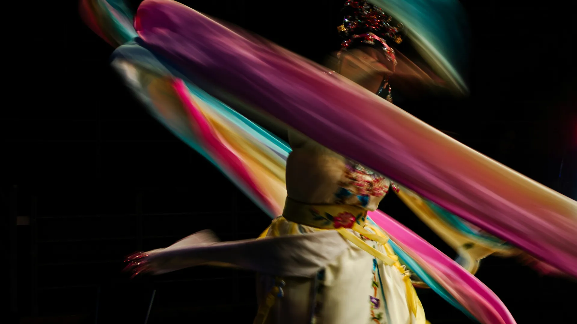 A dramatic photo of a dancer midspin. The colourful ribbons spinning around her body are blurred across her face.